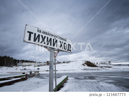Snowy scenery with a Russian sign reading 'Quiet Water'. Icy water and cloudy sky in the background Snowy scenery with a Russian sign reading 'Quiet Water'. Icy water and cloudy sky in the background 121667773