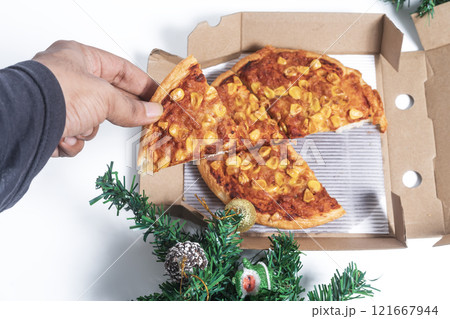 Hand takes a slice of pizza topped with corn from a box amidst Christmas decorations, symbolizing a festive meal 121667944
