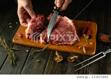 A skilled hand expertly slices raw meat on a rustic wooden cutting board. Surrounding the meat are herbs and spices, enhancing the culinary atmosphere in a cozy kitchen setting 121671602