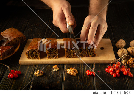 Hands skillfully slice a moist and flavorful cake adorned with nuts and bright red berries. The rustic wooden table provides a charming backdrop, enhancing the delightful atmosphere of the moment 121671608