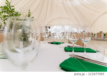 Close up of wine glasses, silver tableware and green napkins on round tables covered with white tablecloth. Close up of wine glasses, silver tableware and green napkins on round tables covered with white tablecloth. 121677995