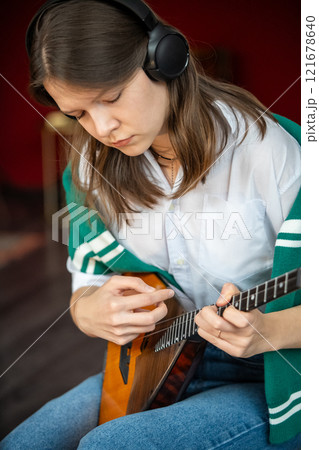 Woman practices balalaika chords wearing headphones while playing in rhythm. Music practice at home 121678640
