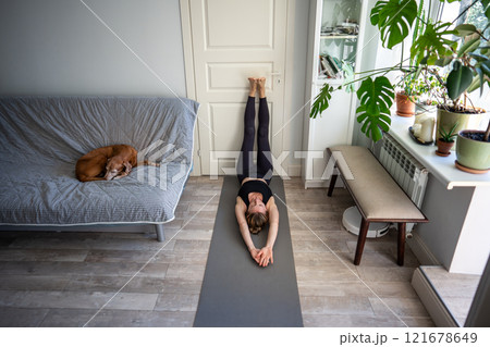 Spiritual woman lies on yoga mat with feet against door, arms stretched above head, dog nap on couch 121678649