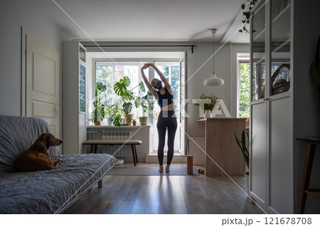 Stretching woman with arms raised above head leans to side on mat while dog watch at owner at home 121678708