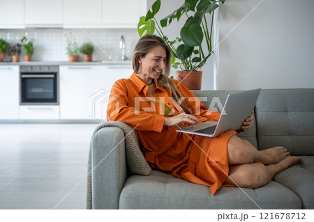 Joyful middle aged woman using laptop smiling relaxing on couch spending leisure time at cozy home. Joyful middle aged woman using laptop smiling relaxing on couch spending leisure time at cozy home. 121678712