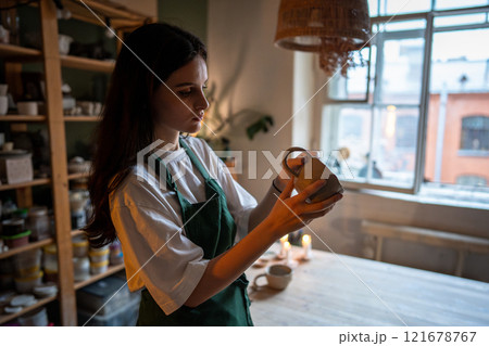 Female artisan examining freshly made clay cup, evaluating form and texture in pottery studio class 121678767