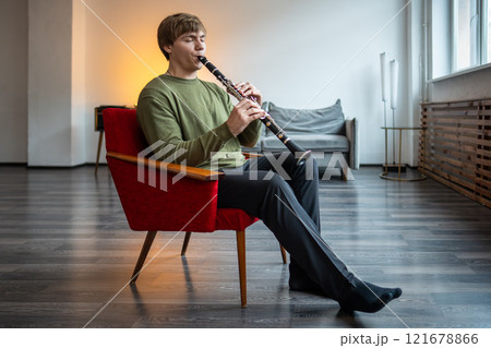 Clarinet player. Man clarinetist playing flute woodwind music instrument sitting on armchair at home 121678866