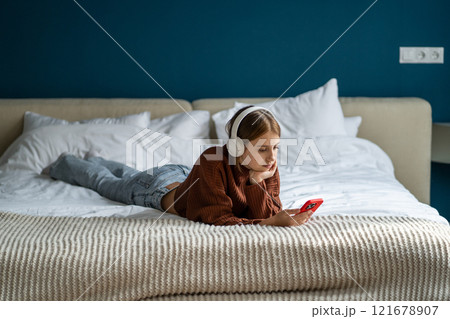 Pensive teenage girl wearing headphones listens to podcast on smartphone at home while lying on bed 121678907