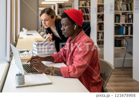 Interested african american student typing on laptop near focused college teacher looking at screen 121678908
