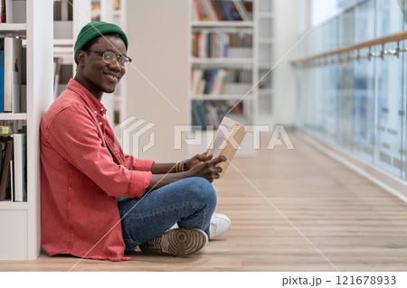 Smiling Black student man reading book in university library , looking at camera. Reading hobby.  121678933
