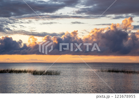 Calm lakeside at sunset in summer evening. Grassy seashore in evening against a cloudy sunset sky. 121678955