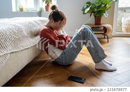 Unhappy teen girl covering face with hands crying while sitting on floor with mobile phone nearby 121678977