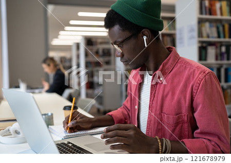 Focused emotionless African American man making notes in notebook sits at table with laptop 121678979