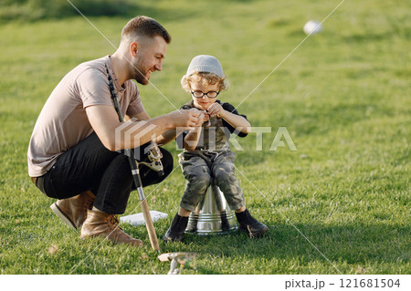 Dad and his child boy spend time outdoors together. Curly toddler boy wearing a khaki overall. Boy sitting on a bucket and listening his father. Dad and his child boy spend time outdoors together. Curly toddler boy wearing a khaki overall. Boy sitting on a bucket and listening his father. 121681504