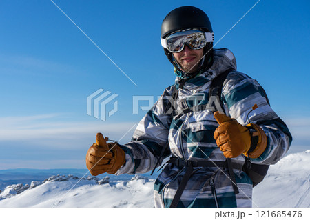Smiling man in skiing or snowboarding winter equipment shows thumbs up. Warm jacket, brown gloves, ski goggles, helmet 121685476