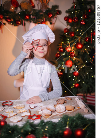 Cheerful girl in chef's hat shows looking through baking pan of cookies on Christmas background Cheerful girl in chef's hat shows looking through baking pan of cookies on Christmas background 121689767