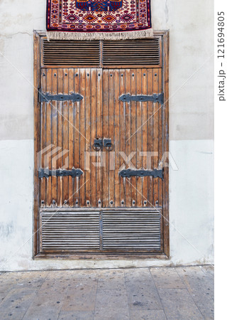 Weathered wooden double doors with metal accents stand against a whitewashed wall. A colorful rug hangs above Weathered wooden double doors with metal accents stand against a whitewashed wall. A colorful rug hangs above 121694805