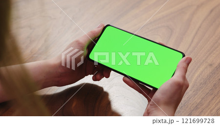 Young woman sitting at a table and using a smartphone with horizontal green screen 121699728