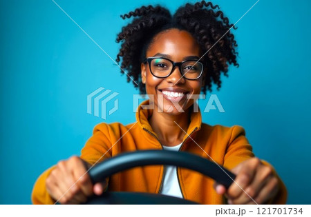 Young beautiful Black curly Female in glasses, hand holds a steering wheel, smiling. Portrait of happy young african american woman on a blue background. Driving lessons and transport industry 121701734