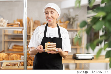 Mature saleswoman offering a piece of cake in plate in bakery 121707057