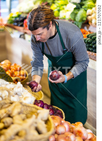 Male grocery store worker arranges red onion and other vegetables on the counter 121707082