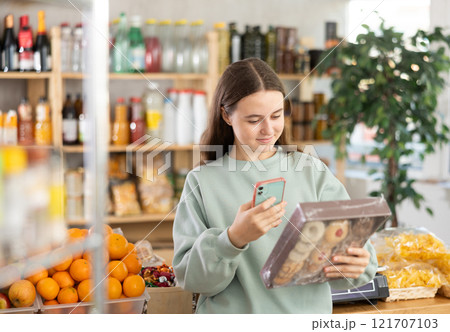 European teen girl customer at shop scans QR code on cookies package using phone camera 121707103