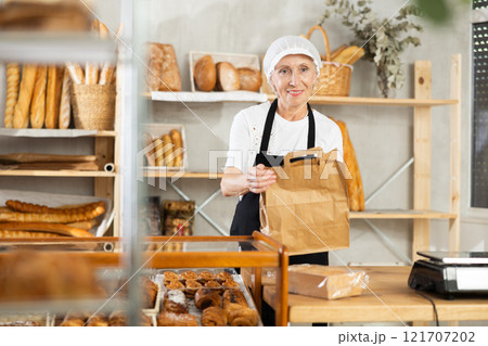 Portrait of helpful female baker with paper bag of fresh baked goods in interior of bakery 121707202