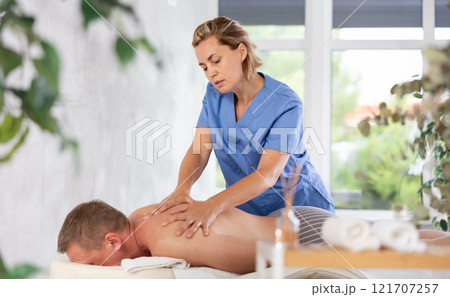 Female masseur performs back stroking and patting during massage session for male client 121707257