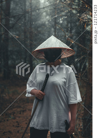 A mysterious man in a conical hat and mask stands in the forest holding a katana. The man is dressed in a white shirt and black pants. The warrior practices with a katana in the rain. 121714233