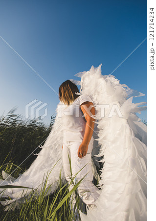 An ethereal angel stands against a blue sky. A woman with large white angel wings stands in a field of tall grass, against a clear blue sky, creating a serene and ethereal atmosphere. An ethereal angel stands against a blue sky. A woman with large white angel wings stands in a field of tall grass, against a clear blue sky, creating a serene and ethereal atmosphere. 121714234