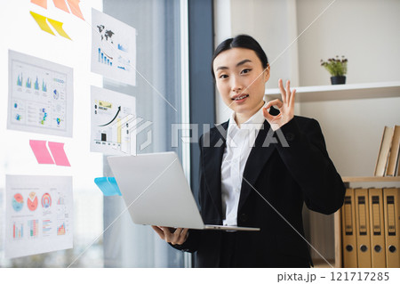 Asian businesswoman in office, holding laptop, presenting data. Young woman in professional attire, showing confidence and success in business environment. Charts and graphs visible on wall. Asian businesswoman in office, holding laptop, presenting data. Young woman in professional attire, showing confidence and success in business environment. Charts and graphs visible on wall. 121717285