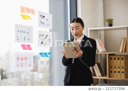 Asian businesswoman in formal attire analyzing data charts on digital tablet in modern office. Reflective charts on window indicate strategic planning and productivity. 121717306