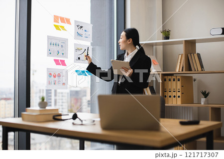 Young Asian businesswoman in office analyzes data charts on glass wall. She uses tablet for insights, exemplifying modern work. Professional setting with city view enhances focus. 121717307