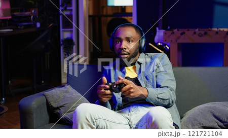 Enthusiastic gamer on couch using controller and headphones to play videogame in neon lit apartment. Player in RGB illuminated living room using high tech gaming console joystick, camera A 121721104