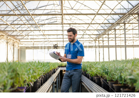 Young bearded man in a blue thisrt standing in a greenhouse and looking enjoyed 121721426