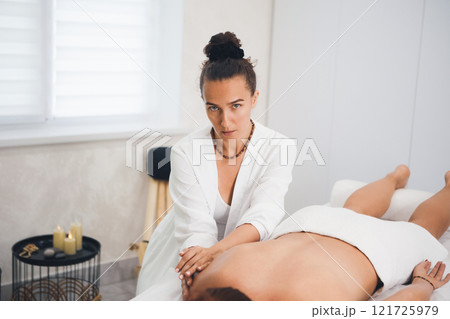 Attractive masseuse in white outfit gently massages a lying woman in pro salon, copy space, selective focus. Attractive masseuse in white outfit gently massages a lying woman in pro salon, copy space, selective focus. 121725979