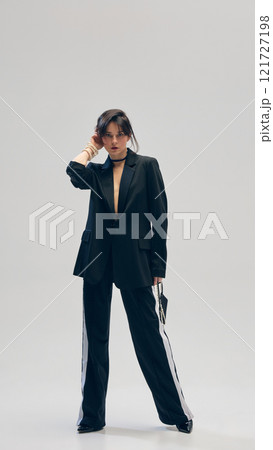 Full-length portrait of young woman wears sport style pants and oversized black blazer with golden accessories, posing against white studio background Full-length portrait of young woman wears sport style pants and oversized black blazer with golden accessories, posing against white studio background 121727198