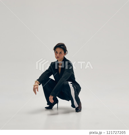 Attractive young woman wears heeled shoes, sport style pants and oversized black blazer and posing sitting against white studio background 121727205
