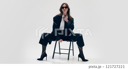 Modern business trends in fashion. Confident young woman poses on chair in black suit with white shirt, tie, glasses and heels, against studio background Modern business trends in fashion. Confident young woman poses on chair in black suit with white shirt, tie, glasses and heels, against studio background 121727221