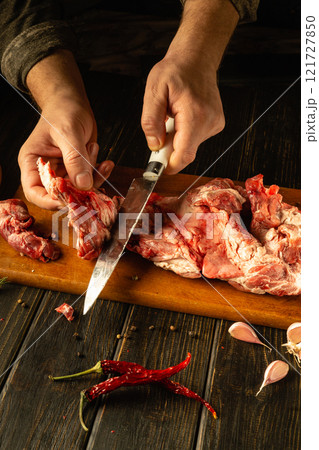 Hands skillfully cut raw lamb meat on a wooden board, surrounded by vibrant spices and garlic. The warm glow of the rustic kitchen enhances the preparation of a hearty dish 121727850