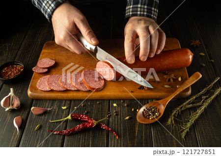 Hands skillfully slice into a rich sausage on a wooden board, surrounded by fresh garlic, herbs, and vibrant spices, evoking the essence of culinary creativity in a cozy kitchen setting 121727851