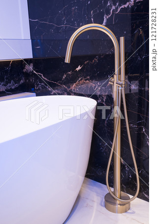 Long stylish Gold faucet on the floor near white bathtub on granite wall background. Modern bathroom interior 121728231