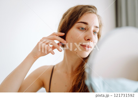 Curly woman touches red inflammations on her face while looking in the mirror in a bright room. Curly woman touches red inflammations on her face while looking in the mirror in a bright room. 121731592
