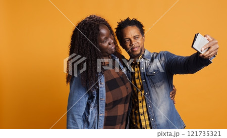 African american husband and wife having fun taking selfies together on phone against yellow background, enjoying leisure time. Joyful confident married people laughing for pictures. Camera B. African american husband and wife having fun taking selfies together on phone against yellow background, enjoying leisure time. Joyful confident married people laughing for pictures. Camera B. 121735321