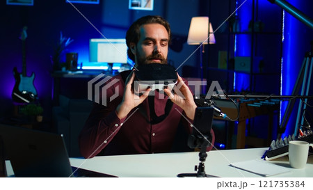 Media star in neon lit studio presents VR goggles from brand sponsoring video, closely analyzing features. Influencer reviewing virtual reality headset for tech enthusiasts watching, camera B Media star in neon lit studio presents VR goggles from brand sponsoring video, closely analyzing features. Influencer reviewing virtual reality headset for tech enthusiasts watching, camera B 121735384
