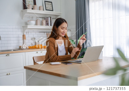 Beautiful young Asian woman working with laptop computer in home kitchen. Work at home 121737355