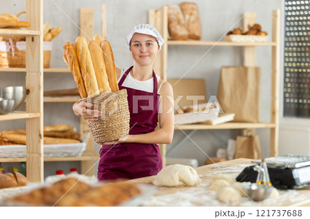 Young girl baker are standing near product range in bakery kitchen with basket loaf of baguettes Young girl baker are standing near product range in bakery kitchen with basket loaf of baguettes 121737688
