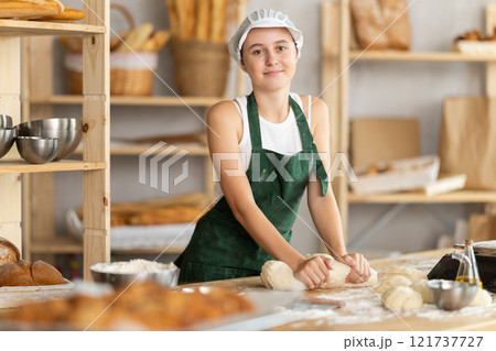 Teen girl works in bakery as baker, kneads dough, works with flour. 121737727
