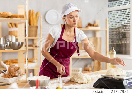Female baker cutting raw dough with knife and weighs pieces of dough on scales for making croissants 121737986