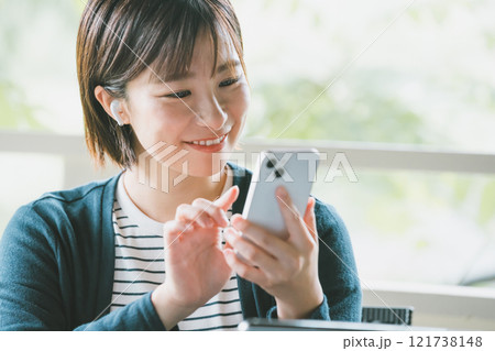緑豊かなベランダでワイヤレスイヤホンをつけてスマホを操作する若い女性 緑豊かなベランダでワイヤレスイヤホンをつけてスマホを操作する若い女性 121738148